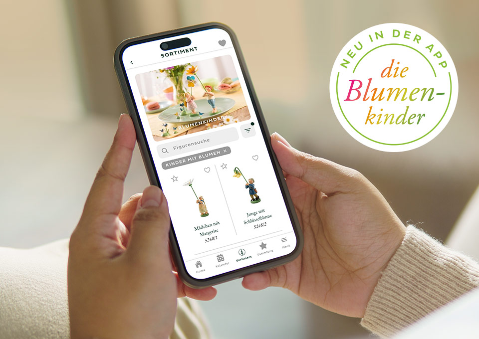 Smartphone mit geöffneter App von Wendt und Kühn, die Blumenkinder-Figuren zeigt, gehalten von zwei Händen vor hellem Hintergrund